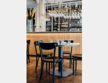 Modernes Restaurantinterieur mit einem runden Tisch aus Marmor und schwarzen Holzstühlen auf einem Holzboden. Im Hintergrund eine beleuchtete Bar mit hängenden Weingläsern und Regalen voller Flaschen.