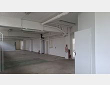 Großer, leerer Innenraum in einem Industriegebäude mit weiß gestrichenen Wänden und Decke, sichtbaren Rohrleitungen und Leuchtstofflampen. Betonboden, mehrere Durchgänge und Türen sowie ein Feuerlöscher an der Wand sind zu sehen.