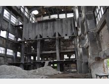 Verlassene Industriehalle mit hohen, beschädigten Betonwänden und großen, teilweise zerstörten Fenstern. Im Inneren befinden sich massive Stahlkonstruktionen und Schutthaufen auf dem Boden, während die Decken und Wände sichtbare Verfallsspuren zeigen.