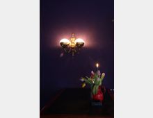 Ein dunkler Raum mit einer violetten Wand, an der eine antike, goldfarbene Wandleuchte mit drei Lampen angebracht ist. Auf einem dunklen Tisch steht eine rote Kanne mit bunten Tulpen und einer brennenden Kerze.
