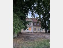 Ein zweistöckiges historisches Gebäude mit beigem Putz und roten Dachziegeln, teilweise verdeckt von großen Bäumen. Die Fassade zeigt mehrere Sprossenfenster, eine mittig platzierte Eingangstür ist von einem angestrahlten Innenraum sichtbar.