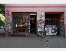 Fassade eines Gebäudes mit einem Glasfenster, das ein Fotostudio zeigt, umgeben von Wänden mit bunten Graffiti und Plakaten. Rechts befinden sich ein Gittertor und eine mit Aufklebern bedeckte Fläche, der Gehweg besteht aus Kopfsteinpflaster.