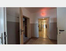 Flur in einem Gefängnis oder einer Haftanstalt mit offenen Türen auf beiden Seiten. Am Ende des Ganges sind vergitterte Türen und eine aufleuchtende rote Lampe über einem Türrahmen zu sehen, der Boden ist mit einem gemusterten Linoleumbelag versehen.