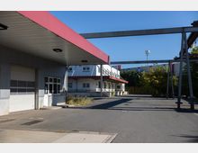 Industriegelände mit mehreren Gebäuden aus grauem und weißem Blech mit roten Akzenten, Überdachungen sowie Stahlträgern im Vordergrund. Der Bereich umfasst eine asphaltierte Fläche und ist von Bäumen und Sträuchern eingefasst.