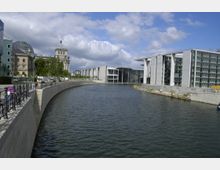 Flussufer in Berlin mit Blick auf moderne Regierungsgebäude und den Reichstag im Hintergrund. Links sind Geländer entlang einer Uferpromenade zu sehen, während rechts der Fluss von klaren, geometrischen Bauten eingefasst wird.