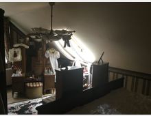 Ein Zimmer mit Dachschräge, möbliert mit antiken Holzmöbeln wie einem Schminktisch mit großem Spiegel, verzierten Stühlen und einer Kommode. Das Licht fällt durch ein großes, stoffverziertes Fenster und betont weiße Kleider und Dekorationsgegenstände.