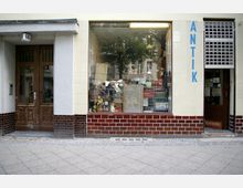 Fassade eines Geschäfts mit der Aufschrift "ANTIK", bestehend aus einem großen Schaufenster voller Antiquitäten und einem offenen Eingang rechts. Links daneben eine hölzerne Tür mit der Hausnummer 66, umgeben von cremefarbenen und braunen Wandfliesen.