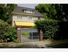 Zweistöckiges Einfamilienhaus mit hellem Putz, dunklem Walmdach und gelber Markise über einem Balkon. Das Grundstück ist von dichten Hecken umgeben und mit einem metallenen Tor sowie Backsteinsäulen abgegrenzt.