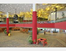 Großes Foyer eines Veranstaltungsgebäudes mit rotem Geländer im Obergeschoss und roten Vorhängen an Säulen. Die Decke ist mit goldfarbenen geometrischen Elementen gestaltet, und im Raum sind Pflanzen, verschiedene Sitzbereiche und eine Theke sichtbar.