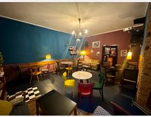 Gemütlicher Lounge- oder Caféraum mit blau- und bordeauxfarbenen Wänden, gemischten kleinen Tischen und Stühlen sowie Sofas und Stehlampen. An der Decke hängt ein Kronleuchter, an der Rückwand stehen Regale und gerahmte Bilder, daneben eine Tür mit Plakaten.