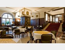 Innenraum eines eleganten Restaurants mit weißen Tischdecken, dunklen Holzmöbeln und goldenen Verzierungen. Hohe Fenster lassen Tageslicht herein, während eine rote Teppichtreppe zu einem erhöhten Bereich führt.