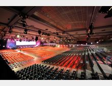 Großer Veranstaltungssaal mit einer Bühne im Hintergrund, umgeben von zahlreichen Scheinwerfern und technischen Geräten. Reihen von Stühlen in verschiedenen Farben füllen den Raum, der durch eine hohe Decke mit einem Gittermuster und Beleuchtung ausgestattet ist.