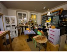 Gemütlicher Innenraum einer kleinen Bar oder eines Cafés mit Holzboden, kleinen Tischen und Stühlen sowie einer Theke mit Flaschenregalen. Im Hintergrund sind zwei Fenster, eine Glastür und ein Getränkekühlschrank zu sehen, daneben gestapelte Kisten und Dekoration auf Regalen.