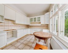 Helle Küche mit weißen Einbauschränken und beige gefliesten Wänden sowie einem hexagonalen Fliesenboden; im Vordergrund ein runder Holztisch und große Fensterfront mit Blick ins Grüne.