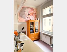 Kleine Küche mit einem hellen Fenster, einer Arbeitsfläche mit Spüle und Kaffeemaschine sowie einem rustikalen Holzvitrinenschrank. An der Wand hängt ein großformatiges Bild mit abstraktem, farbenfrohem Motiv.