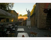 Ein gepflasterter Innenhof mit Tischen und Stühlen eines Biergartens, umgeben von historischen Backsteinmauern und Gebäuden. Im Hintergrund steht eine weiße Statue auf einem Sockel, dahinter eine mit Bögen verzierte Brücke und Bäume.