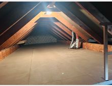 Ausgebauter Dachboden mit schrägen Dachflächen und freiliegenden Holzbalken, beleuchtet durch ein Dachfenster. Der Boden ist mit Platten ausgelegt; an der rechten Seite sind Lüftungsrohre und eine niedrige Abstellfläche zu sehen, im Hintergrund eine Ziegelwand.