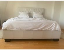 Ein schlichtes Schlafzimmer mit einem großen weißen Polsterbett, das weiße Bettwäsche und zwei Kissen hat. Der Raum hat helle Holzfußböden und eine minimalistische Einrichtung.