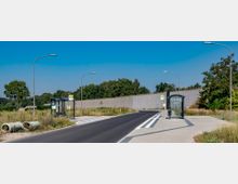 Eine Bushaltestelle an einer asphaltierten Straße mit zwei überdachten Wartehäuschen und Laternenmasten. Im Hintergrund sind eine graue Mauer, Wildwiesen und Bäume unter klarem, blauem Himmel zu sehen.