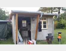 Kleines Gartenhaus aus grauem Putz mit flachem Dach, Regenrinnen und einem offenen Anbau mit Holzstützen; im Inneren sind Leitern und Baumaterial zu sehen. Daneben befindet sich ein Fenster mit hellem Rahmen, umgeben von Rasen und Bäumen.