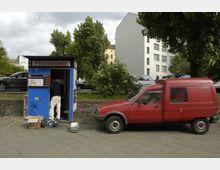 Ein blauer Fotoautomat mit der Aufschrift "Fotoautomat" steht am Straßenrand, daneben ein roter Lieferwagen. Im Hintergrund sind Bäume, geparkte Autos und Gebäude mit hellen Fassaden zu sehen.