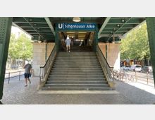 Treppenaufgang zur U-Bahn-Station "Schönhauser Allee" in Berlin, überdacht von einer grün gestrichenen Brückenkonstruktion. Im Hintergrund sind Straßen, parkende Fahrräder und Bäume zu sehen.