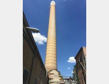 Hoher Industrieschornstein aus gelben Ziegelsteinen, umgeben von Backsteingebäuden eines alten Fabrikgeländes bei klarem, blauem Himmel. Im Vordergrund sind Gebäudefassaden mit Fenstern und Rohrleitungen zu sehen.