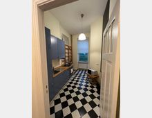 Schmale Küche mit schwarz-weiß kariertem Fliesenboden, blauen Küchenschränken und einer Holzarbeitsplatte. Im Hintergrund ein Fenster mit weißem Rollo, ein Heizkörper darunter sowie ein kleiner Tisch mit Mikrowelle.