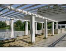 Weiße Pergola mit Säulen und Querbalken neben einem kleinen See, umgeben von Bäumen und Wohngebäuden im Hintergrund. Der Boden ist aus Kies und gepflasterten Flächen, und eine Sitzbank ist unter der Pergola platziert.