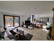 Modernes Wohnzimmer mit offener Gestaltung, großen Fenstern und hellen Fliesenböden. Im Vordergrund befindet sich ein Sofa mit Couchtisch, im Hintergrund eine Terrasse mit Holzüberdachung sowie ein Essbereich mit Tisch und Stühlen.