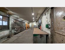 Ein Industrie-innenraum mit abgenutztem Holzboden, rohen Wänden und sichtbaren Rohren sowie elektrischen Leitungen an den Wänden. Links befinden sich große Metallfenster, rechts ein Arbeitstisch und ein Feuerlöscher.