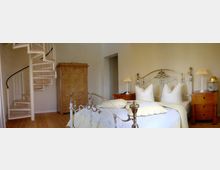 Ein helles Schlafzimmer mit einem weißen Metallbett und verschnörkelten Verzierungen, flankiert von zwei hölzernen Nachttischen mit Lampen. Im Hintergrund stehen eine geschwungene Wendeltreppe aus Holz und Metall sowie ein großer, antik wirkender Kleiderschrank.