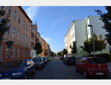 Eine gepflasterte Wohnstraße mit mehrstöckigen Gebäuden auf beiden Seiten, die in verschiedenen Farben gestrichen sind (orange, gelb, rot und weiß). Am Straßenrand sind parkende Autos und einzelne Bäume entlang des Gehwegs zu sehen, bei klarem Himmel und Tageslicht.