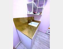 Kleine Küche mit einer rosa Wand, einer Arbeitsplatte aus goldfarbenem Metall, einem weißen Spülbecken und einer Darstellung von offenen Regalen, die mit Geschirr und Lebensmitteln gefüllt sind. Der Boden besteht aus hellem Holz.