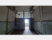 Ein leerer, heruntergekommener Raum mit hellblauen Fliesen an den unteren Wänden und freiliegenden Rohren an der Decke. Im Hintergrund befindet sich eine Tür mit Glasbausteinen darüber, die zu einem helleren Raum führt.