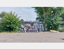 Flacher Platz mit grauen Pflastersteinen, im Hintergrund eine Wand mit großflächigem Graffiti, teilweise von Bäumen und Sträuchern verdeckt. Rechts unten liegen gestapelte Pappkartons, im Hintergrund sind vereinzelte Gebäude erkennbar.