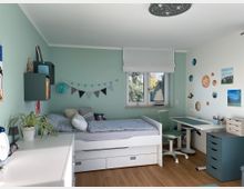 Helles Kinderzimmer mit mintgrünen Wänden, einem weißen Einzelbett mit Schubladen, einem Schreibtisch mit grünem Stuhl und einer Kommode. Die Wand über dem Bett ist mit einer Wimpelkette dekoriert, während die gegenüberliegende Wand ein Planetensystem und Bilder zeigt; ein Fenster mit Blick auf Bäume lässt Tageslicht herein.