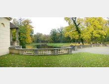 Terrasse mit steinernem Geländer und dekorativen Pflanzgefäßen, angrenzend an eine Rasenfläche und einen Teich. Im Hintergrund herbstliche Bäume in einem Parklandschaft.