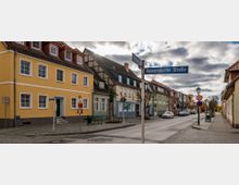 Straßenszene in einer kleinen Stadt mit zweigeschossigen Häusern in verschiedenen Farben, darunter gelbe und grüne Gebäude mit roten Dächern. Im Vordergrund befindet sich ein Straßenschild mit der Beschriftung „Heinersdorfer Straße“ sowie eine Bushaltestelle, Verkehrsschilder und eine gepflasterte Gehwegfläche.