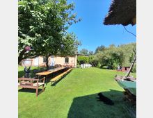 Großer Garten mit Rasenfläche, langen Tischbänken unter einem Baum mit Lampions und einem beige gestrichenen Gebäude im Hintergrund. Im hinteren Bereich stehen weiße Gartenstühle, daneben Bäume und Büsche, sowie ein Holzunterstand und ein Hund im Schatten.