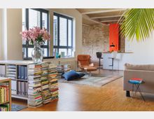 Heller Wohnraum mit modernen und rustikalen Elementen, darunter eine Backsteinwand, große Fenster mit schwarzen Rahmen und Holzboden. Im Vordergrund stehen ein Sessel, ein niedriger Tisch mit Büchern und ein Regal mit gestapelten Büchern, während im Hintergrund eine offene Küche mit Barhockern zu sehen ist.