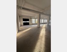 Großer, leerer Industrie- oder Lagerraum mit Betonboden, weißen Stützpfeilern und freiliegenden Deckeninstallationen. An der Seite befinden sich hohe Sprossenfenster und ein großes dunkles Tor, durch die tief stehendes Sonnenlicht lange Schatten wirft.