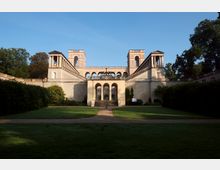 Historisches Gebäude mit symmetrischer Architektur, bestehend aus zwei Türmen, verbunden durch Arkaden und Treppenanlagen links und rechts, umgeben von gepflegtem Rasen und dichter Vegetation. Im Vordergrund führt ein Weg auf das Zentrum des Bauwerks mit einer Veranda mit Rundbögen zu.