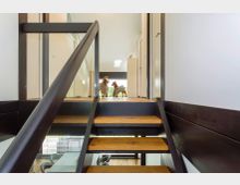 Blick von einer modernen Innentreppe mit offenen Holzstufen und dunklem Metallgeländer in einen hellen Flur mit Glaswänden und Türen. Am Ende des Flurs steht eine große Fensterfront, die Tageslicht hereinlässt und den Blick ins Grüne ermöglicht.