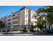 Straßenecke mit einem mehrstöckigen Wohngebäude im klassischen Stil, cremefarbene Fassade mit Balkonen und schrägem Dach. Umgeben von Bäumen, geparkten Autos und einer Ampel; im Vordergrund fährt ein Fahrradfahrer.