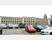 Großer, rechteckiger Schul- oder Verwaltungsbau mit drei Stockwerken, regelmäßiger Fensterfront und beigefarbenen Fassadenfeldern. Davor liegt ein asphaltierter Parkplatz mit mehreren Autos sowie Baustellencontainern und Absperrzäunen; im Hintergrund ragt ein Kirchturm über das Gebäude.