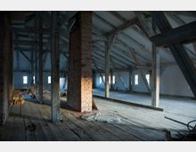 Ein leerer Dachboden mit Holzträgern, einem freistehenden Backsteinschornstein und sichtbaren Holzbalken. Der Boden besteht aus Holzdielen, und kleine Fenster an den Wänden lassen Tageslicht herein.