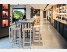 Moderner Weinladen mit langer Regalanlage aus Holz an der rechten Wand, gefüllt mit Weinflaschen, und einer zentralen hohen Tischreihe mit Holz-Hockern. Helle Innenraumbeleuchtung mit Schienenstrahlern, polierter Steinboden und ein Sitzbereich am Fenster mit Blick auf grüne Weinberge.