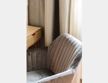 Grauer, gepolsterter Sessel mit vertikalen Nähten neben einem hellen Holztisch mit einer Korbgeflecht-Schublade, vor einem Hintergrund aus grauen Vorhängen und hellem Tageslicht.