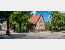 Einfamilienhaus mit rotem Satteldach und verputzter Fassade an einer Ecke einer gepflasterten Straße, umgeben von Bäumen und weiteren Einfamilienhäusern in einer ruhigen Wohngegend. Ein Verkehrsschild und ein kleiner Gartenzaun sind im Vordergrund sichtbar.